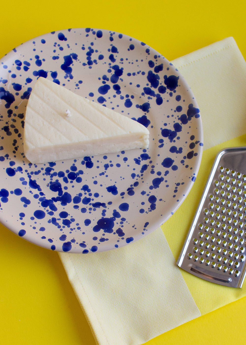 Block of brie cheese on a speckled plate with a metal grater on a yellow background. Fun fact: this is not a real piece of brie, it is a candle! Part of the Playful Food Candle Collection available via Wildschut Antiques Concept Store.