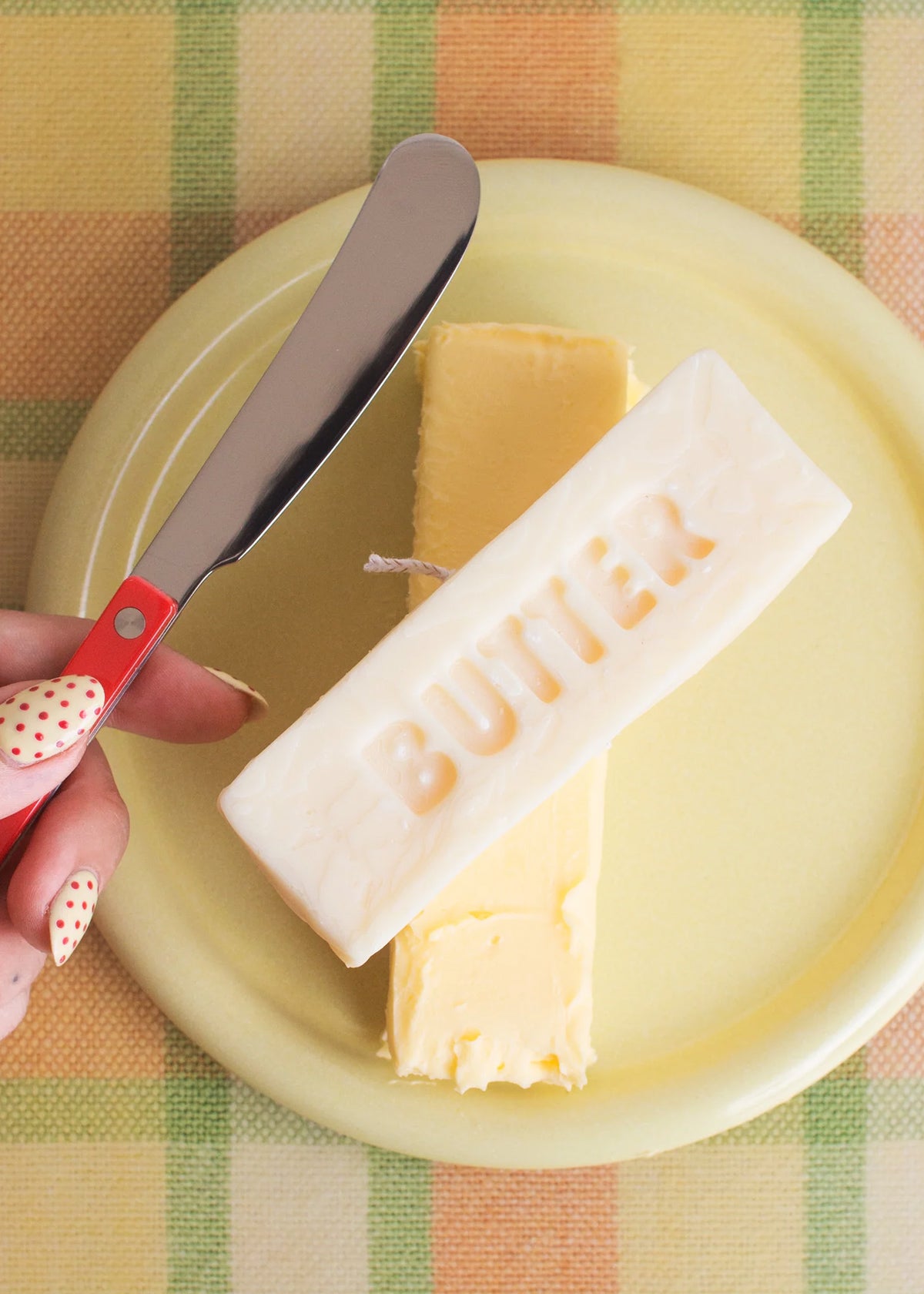 Butter block candle with a knife on a yellow plate against a checkered tablecloth.