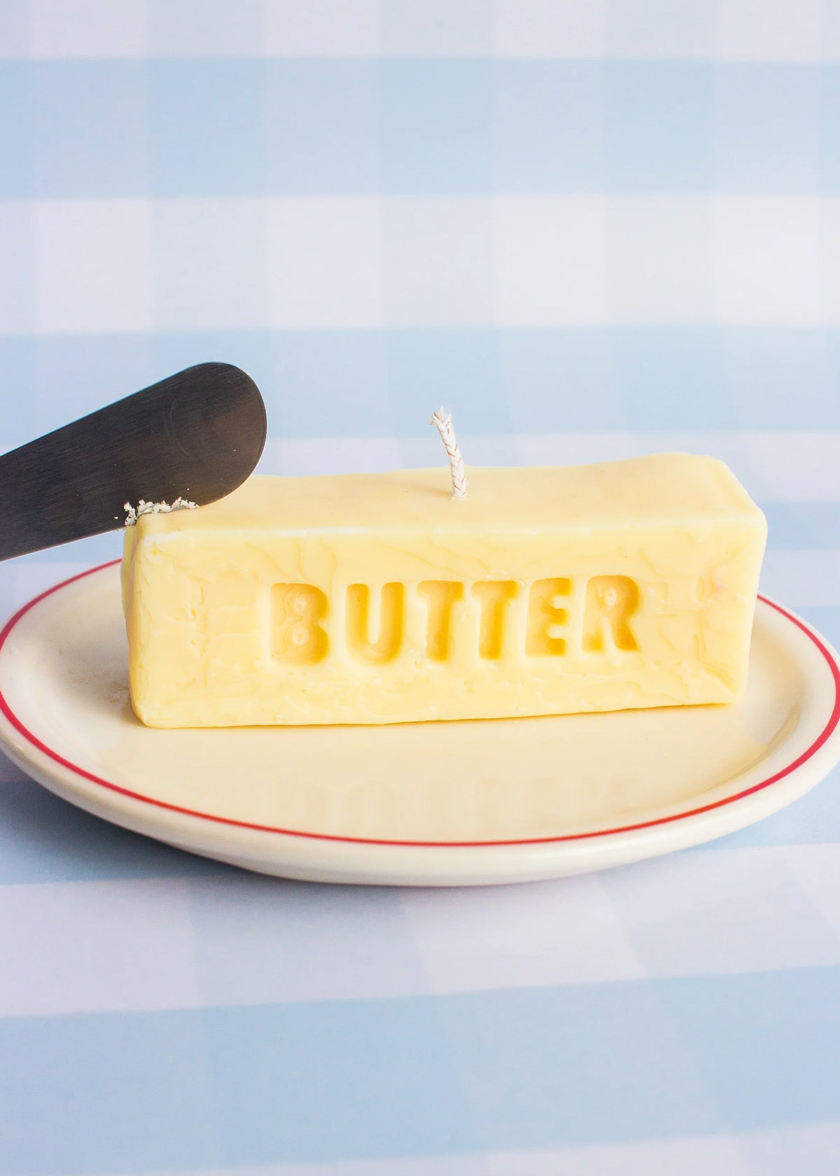 Stick of Butter candle on a plate with a knife against a light blue background. Part of the Playful Food Candle Collection available via Wildschut Antiques Concept Store.