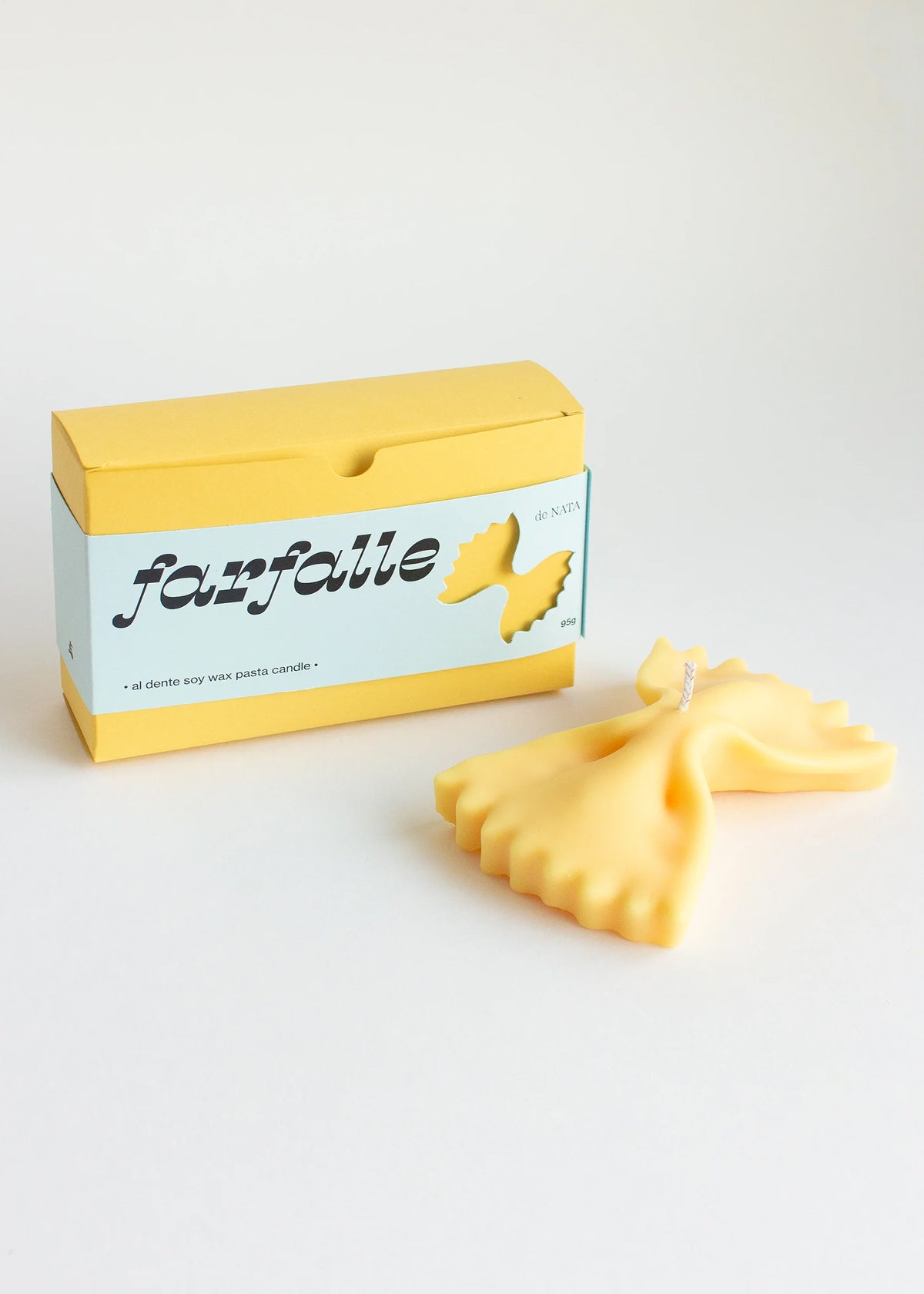 A yellow candle in the shape of a farfalle pasta piece, together with its packaging on a white background. Part of the Playful Food Candle Collection available via Wildschut Antiques Concept Store.