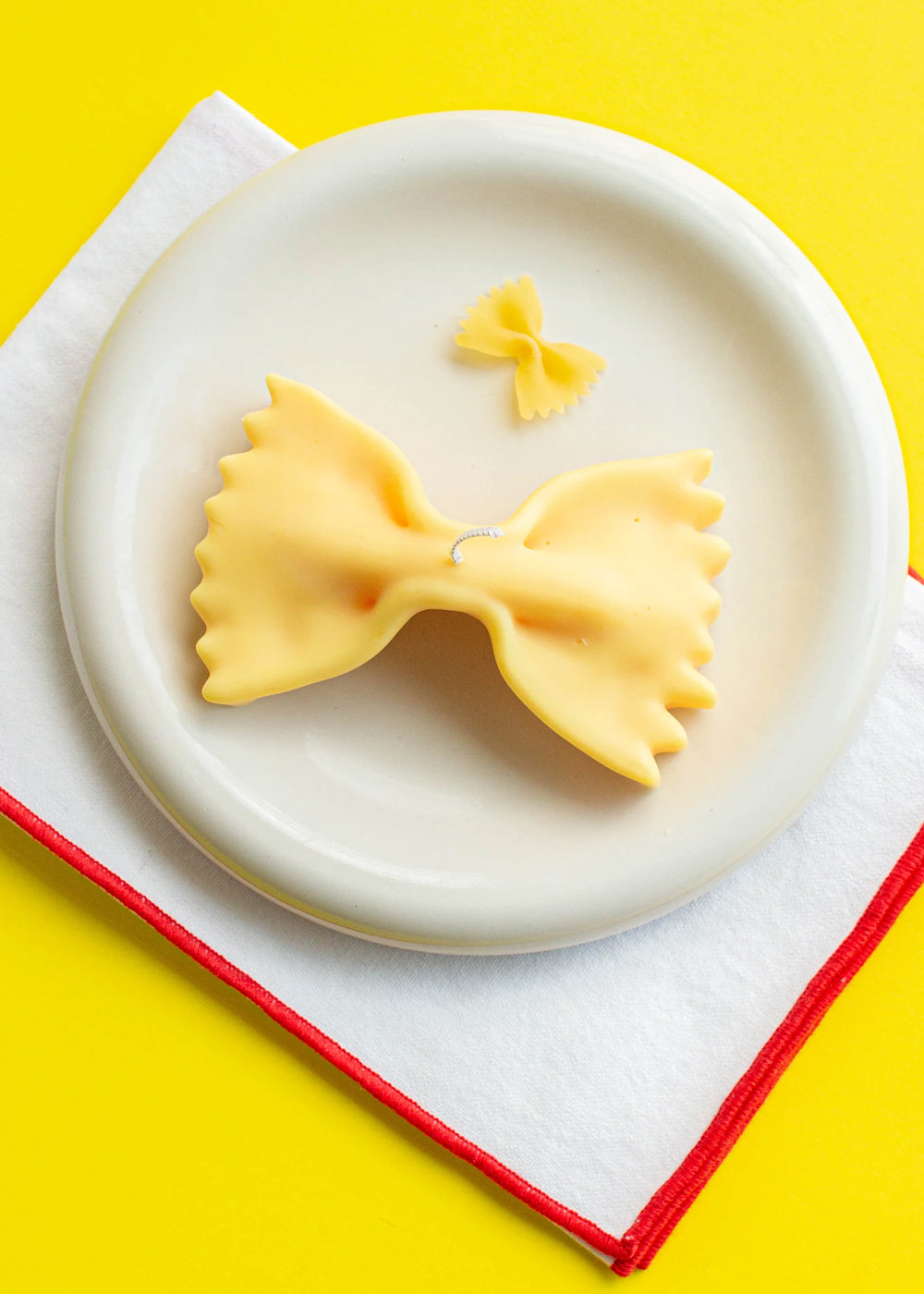 Bow-shaped pasta candle on a white plate with a yellow background. Part of the Playful Food Candle Collection available via Wildschut Antiques Concept Store.