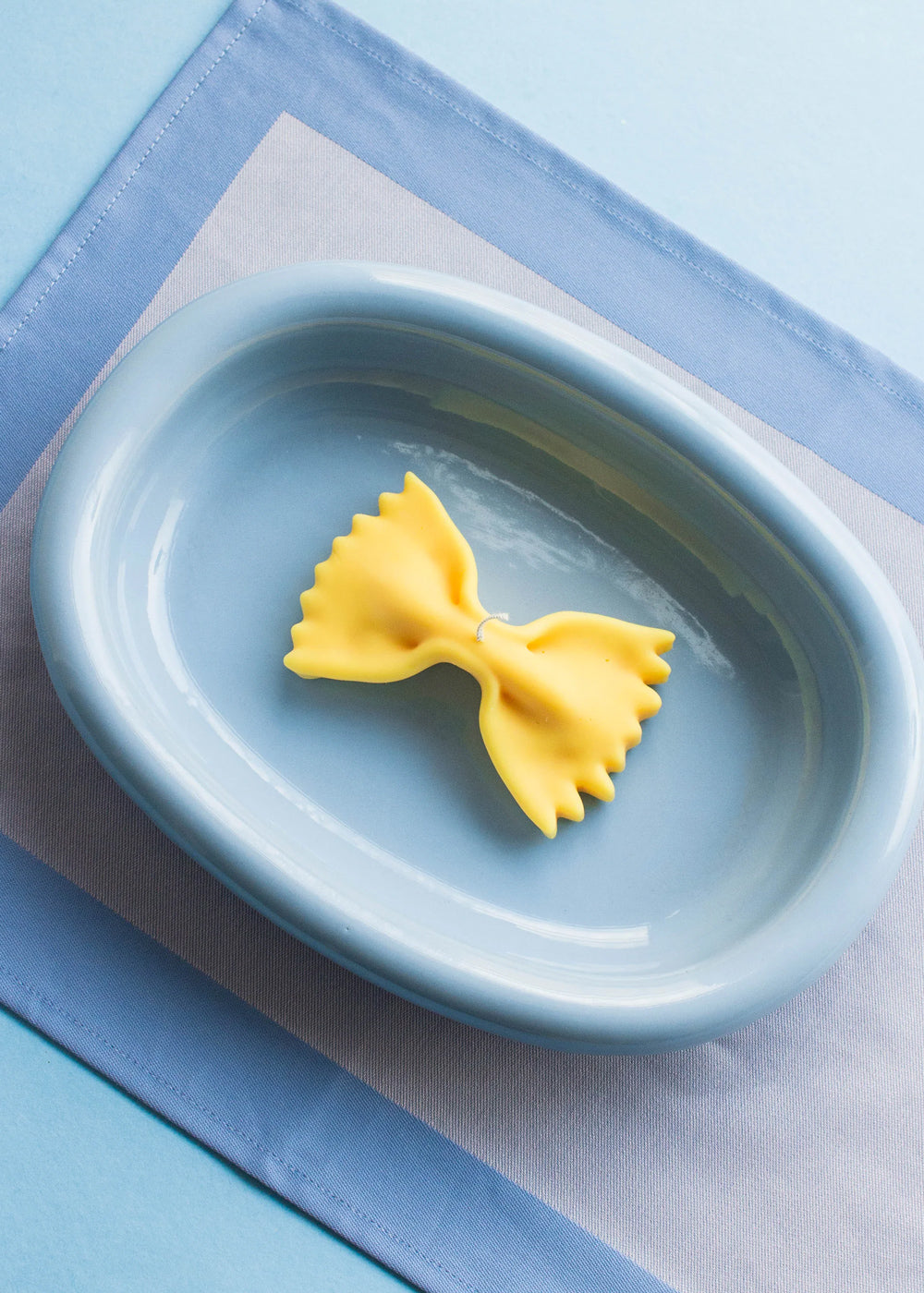 A yellow candle in the shape of a farfalle pasta piece on a blue plate on a blue background. Part of the Playful Food Candle Collection available via Wildschut Antiques Concept Store.