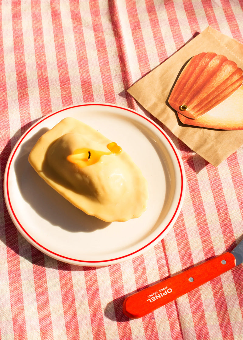 Yellow Madeleine-cake-candle on a white plate with a red rim on a red and white striped tablecloth.