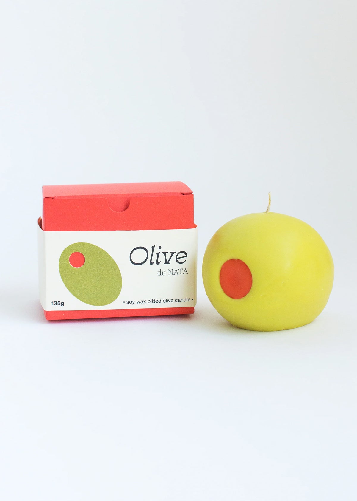 Light green olive-shaped candle with a red center next to its packaging on a white background. Part of the Playful Food Candle Collection available via Wildschut Antiques Concept Store.