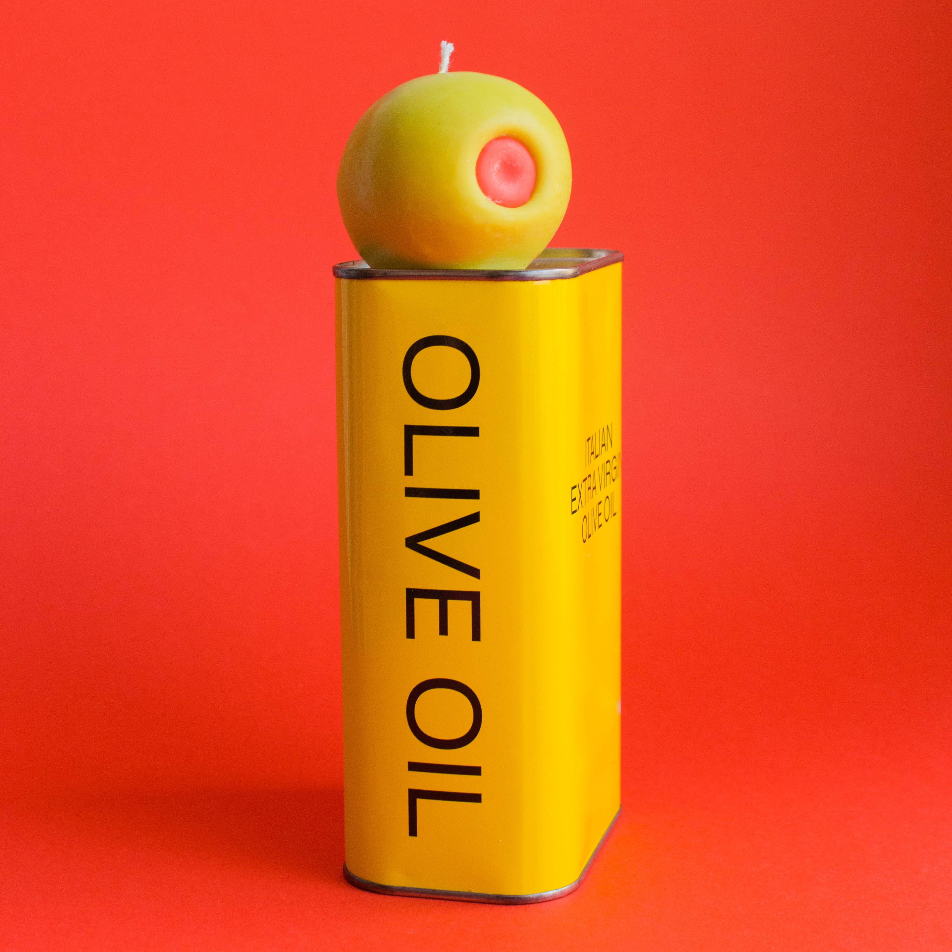 Yellow olive oil can with a green olive candle on top against a red background