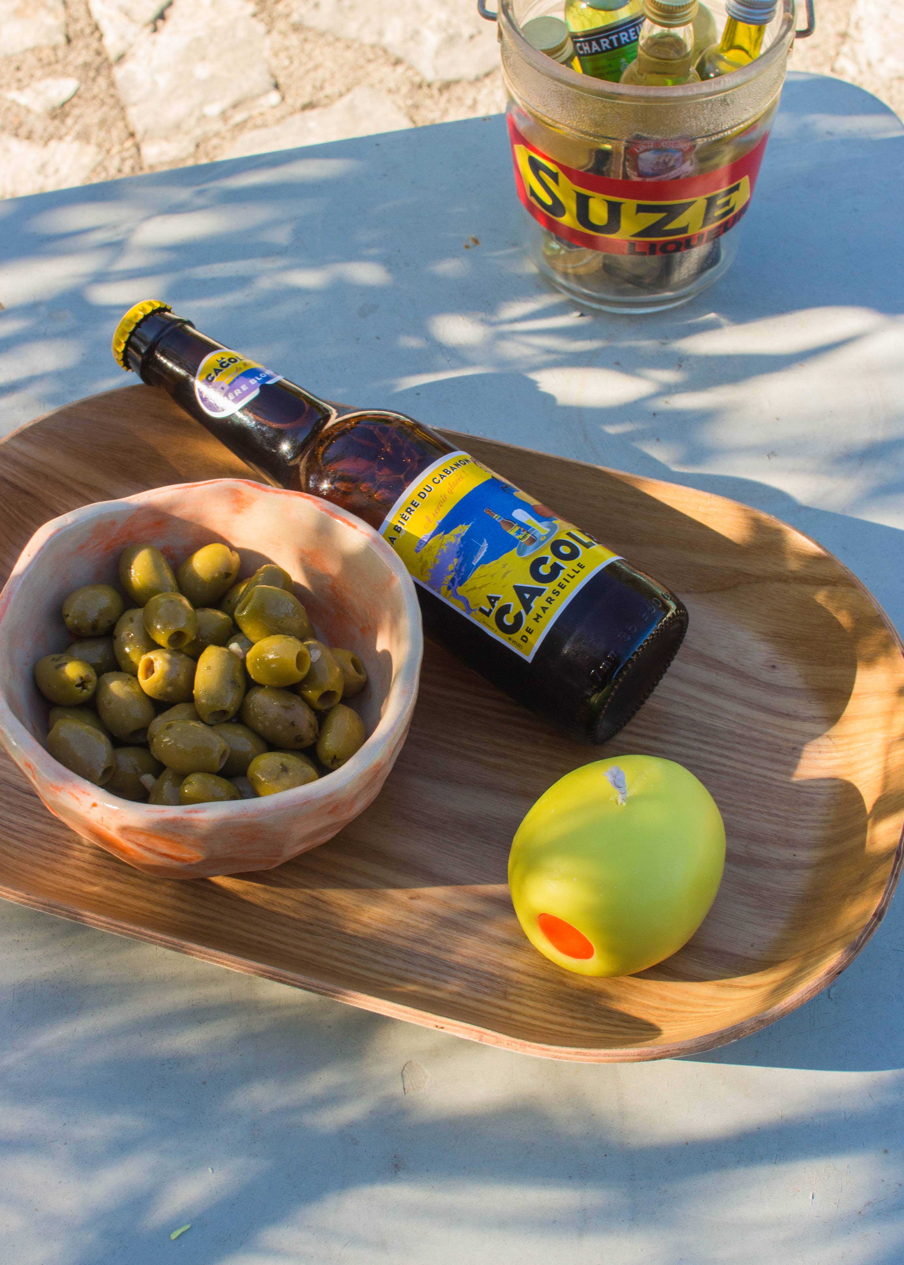 Bottle of beer with a label, bowl of olives, and apple on a green olive candle on a wooden tray outdoors.