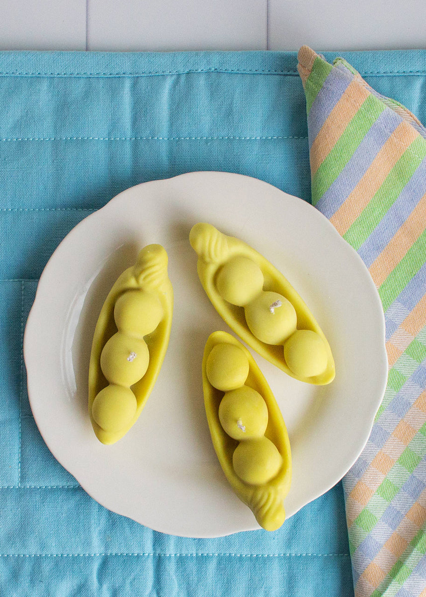 Light green pea pod candles on a white plate with cutlery and a colorful napkin on a blue placemat. Part of the Playful Food Candle Collection available via Wildschut Antiques Concept Store.