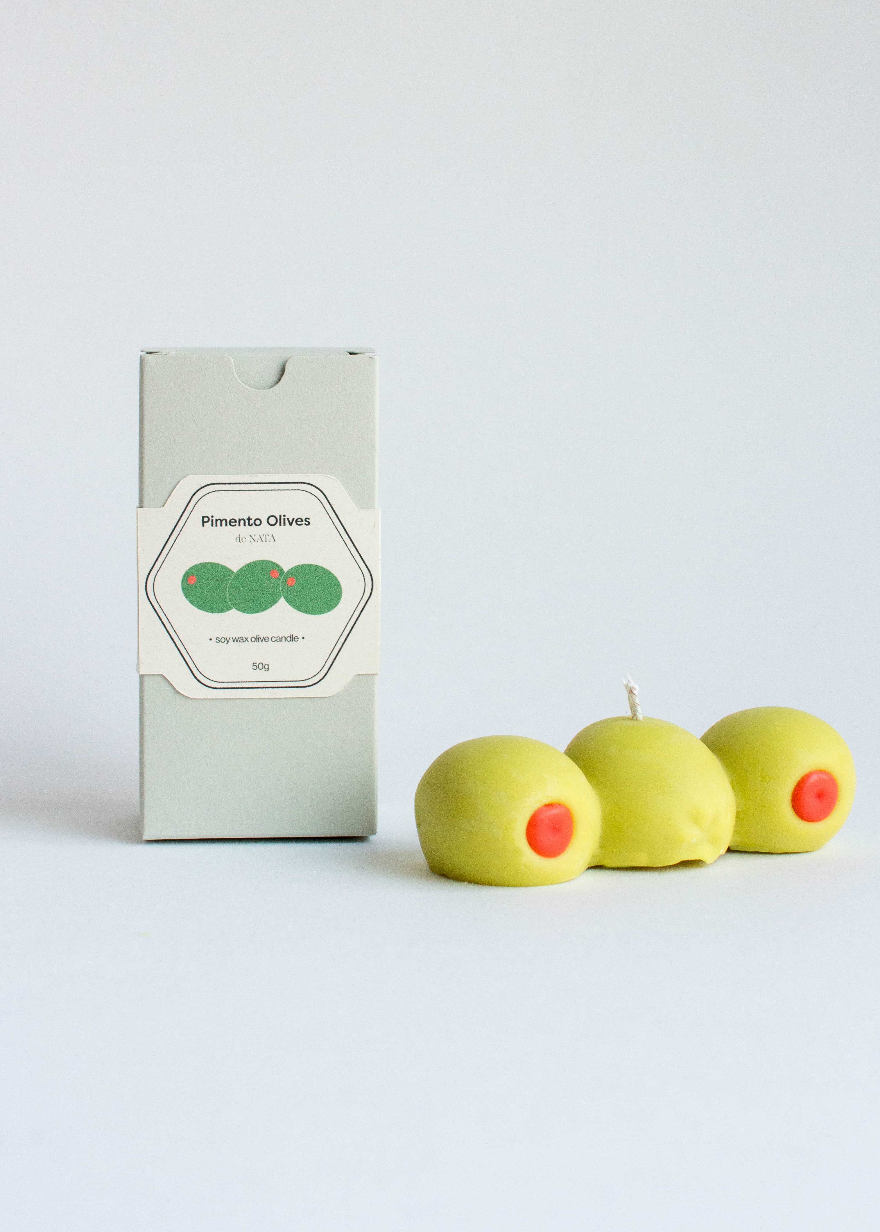 Three green olives candle with red centers and a box labeled 'Pimento Olives' on a white background
