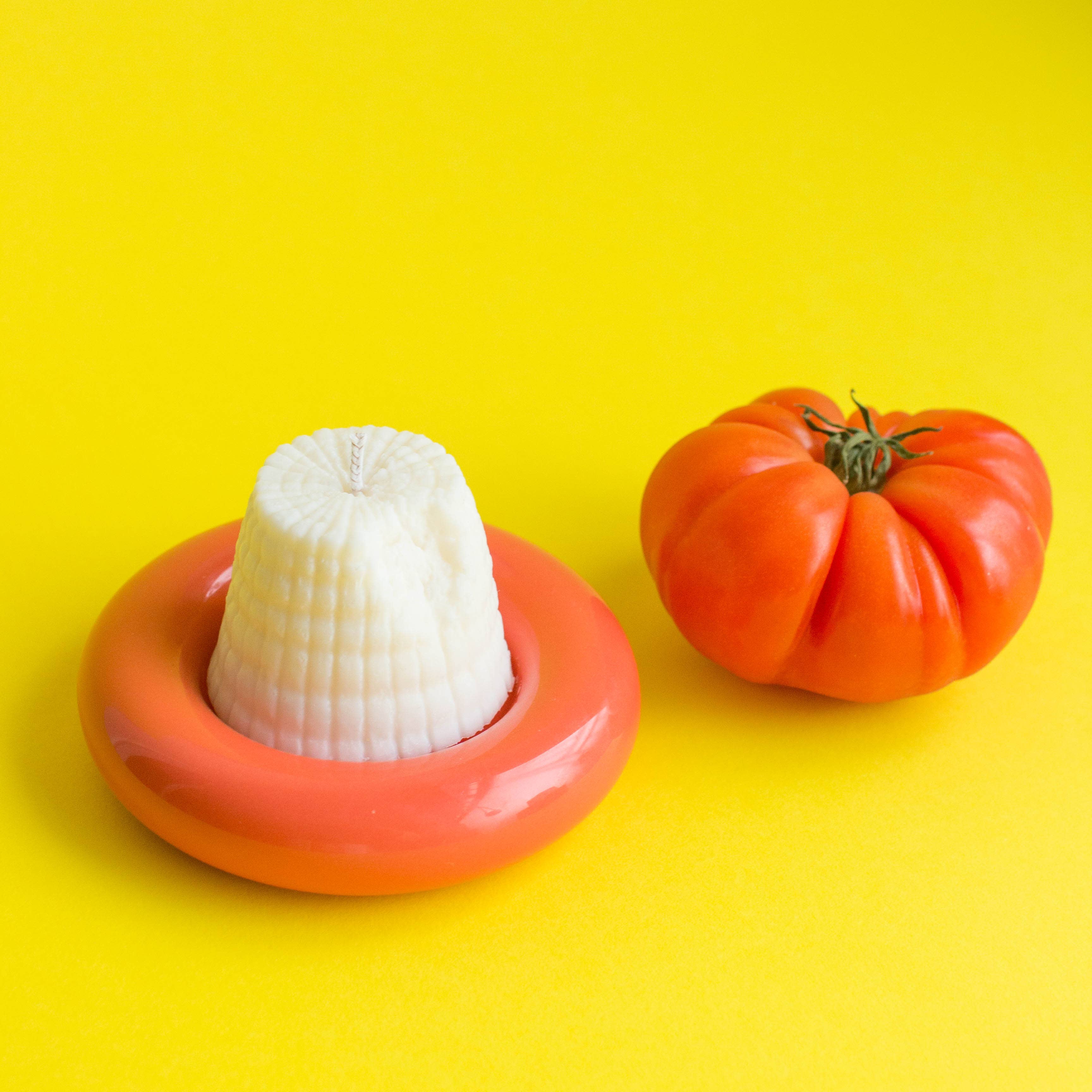 White Candle in the shape of a piece of Ricotta Cheese next to a tomato on a yellow background