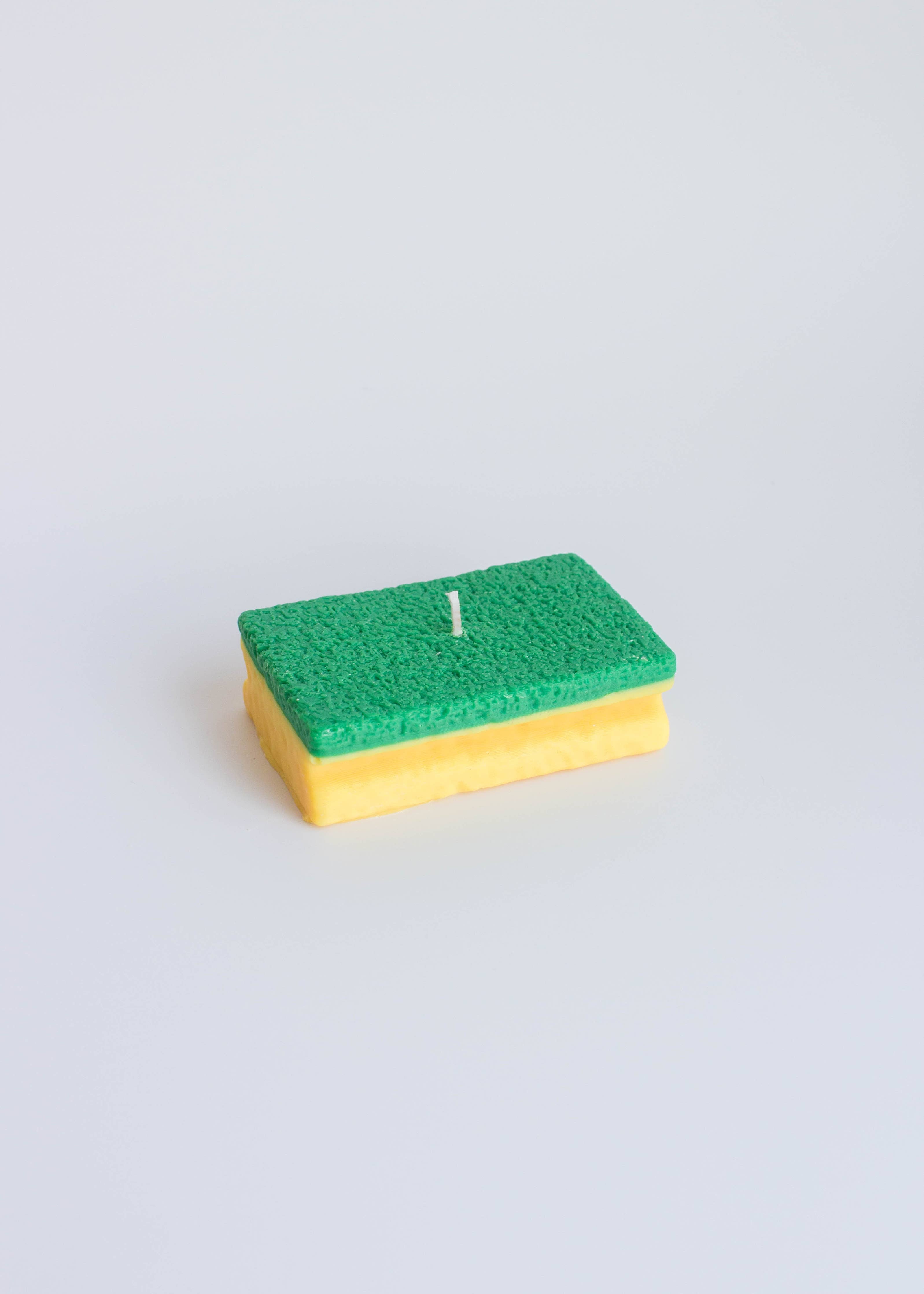 Rectangular candle in the shape of a sponge with green top and yellow bottom on a light gray background, part of the playful food candle collection, available at Wildschut Antiques