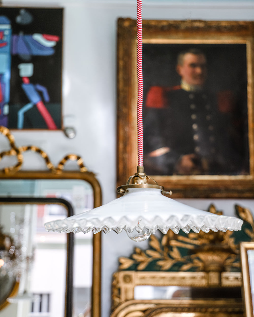 A vintage French white glass frilled coolie pendant light with red and white electrical wiring, hanging from the ceiling in the store of Wildschut Antiques, with antique paintings and mirrors as backdrop
