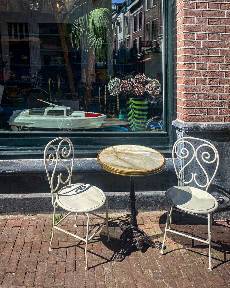 Two white metal chairs and a round table on a brick pavement in front of the store window of Wildschut Antiques in Amsterdam.
