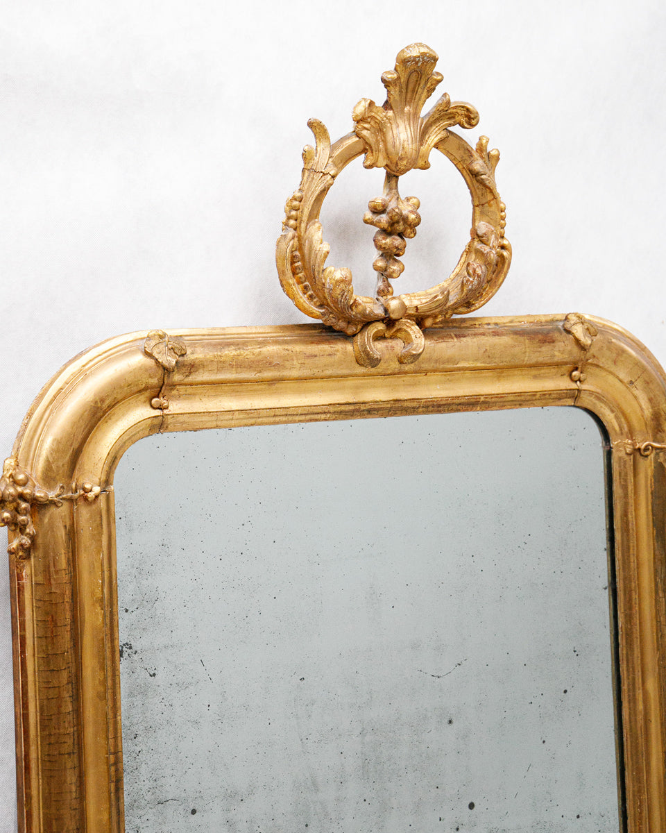 Close-up of a decorative antique French gold-framed mirror with intricate design and grapevine crest on a white background