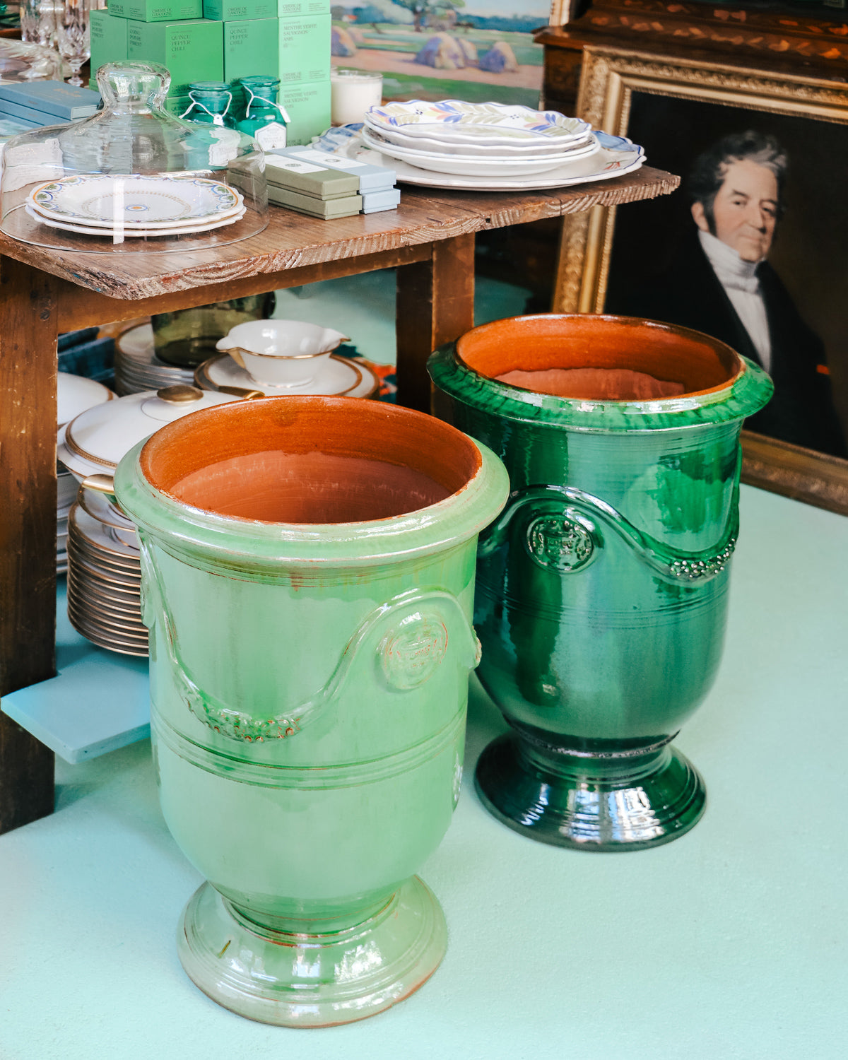 Two large Anduze pots in emerald green and soft jade green, displayed in-store at Wildschut Antiques with an antique portrait painting in the background