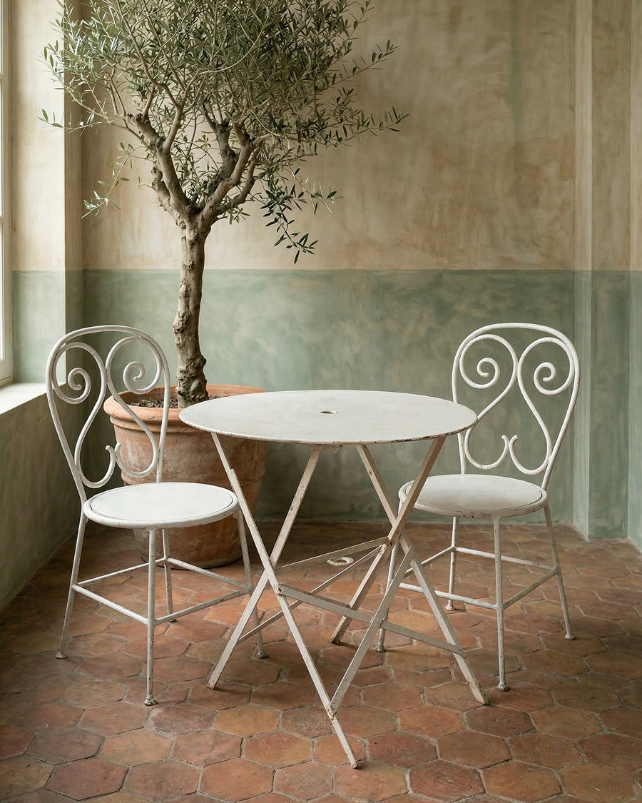Vintage French white metal table and chairs with a potted olive tree against a wall with green and beige tiles, available via Wildschut Antiques.