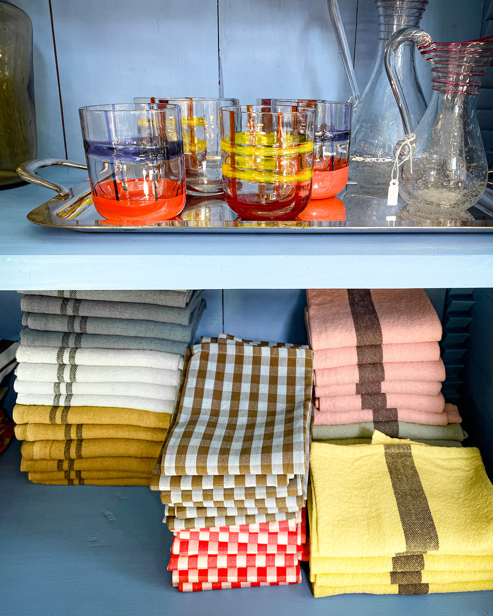 Several piles of neatly folded colorful linen dish towels by Charvet Editions and glasworks by Ichendorf Milano available at Wildschut Antiques