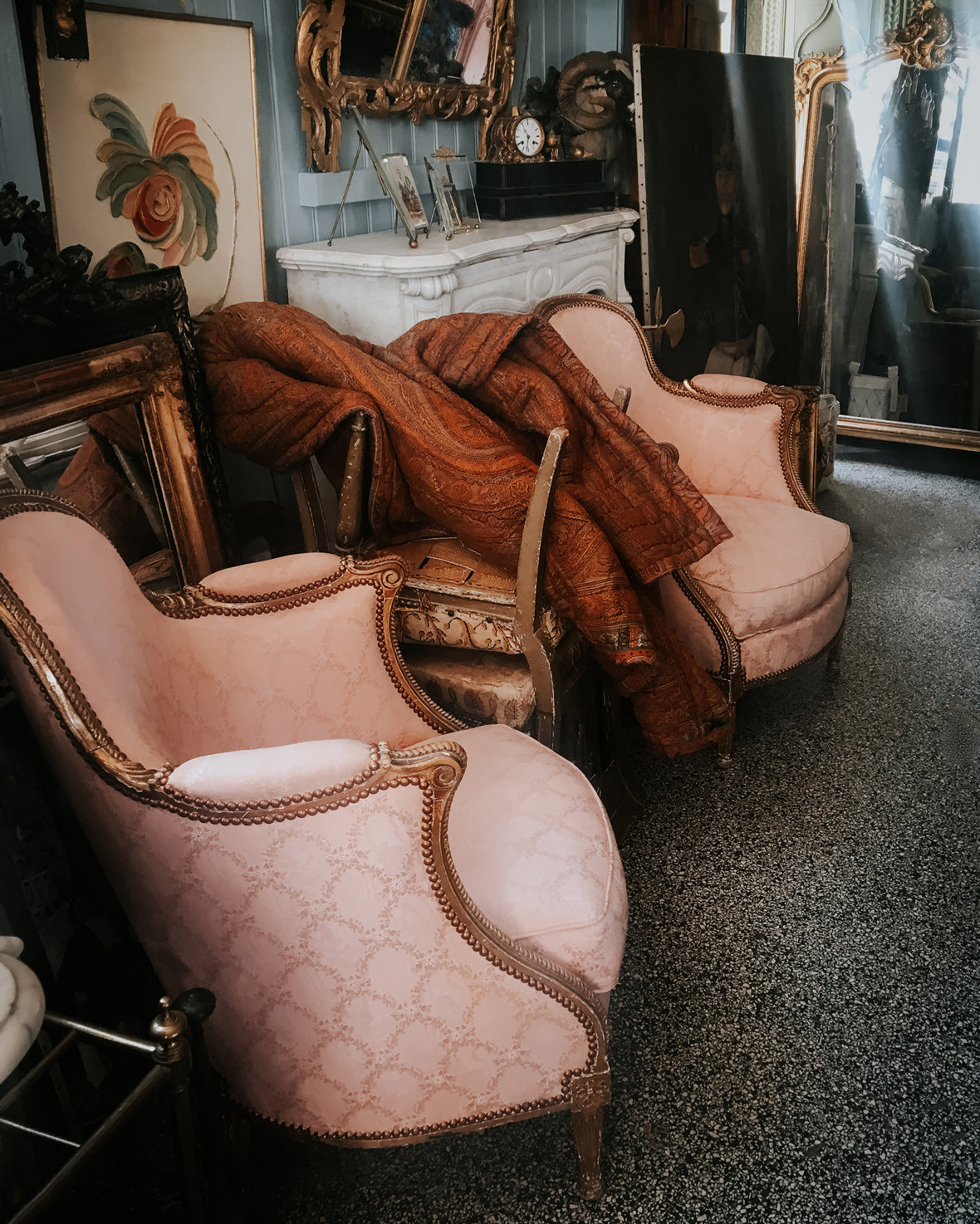 Set of pink bergère chairs with gilded frames, draped with a red wool blanket, positioned beside a white marble fireplace. In the background, antique gilded mirrors and paintings glow in soft, dreamy light—at Wildschut Antiques.