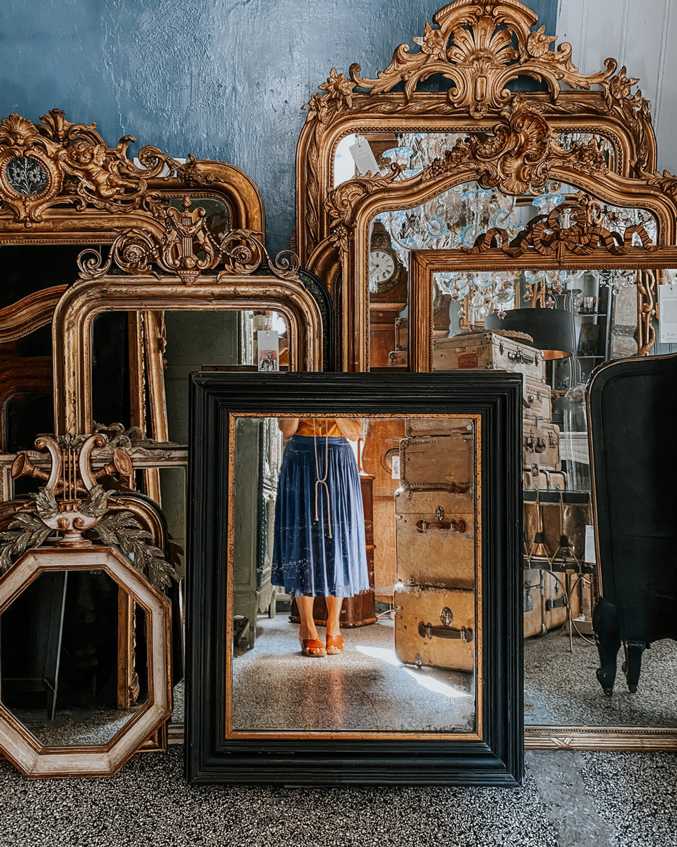 A collection of antique giltwood mirrors set against a wall reflecting a woman in a blue skirt.