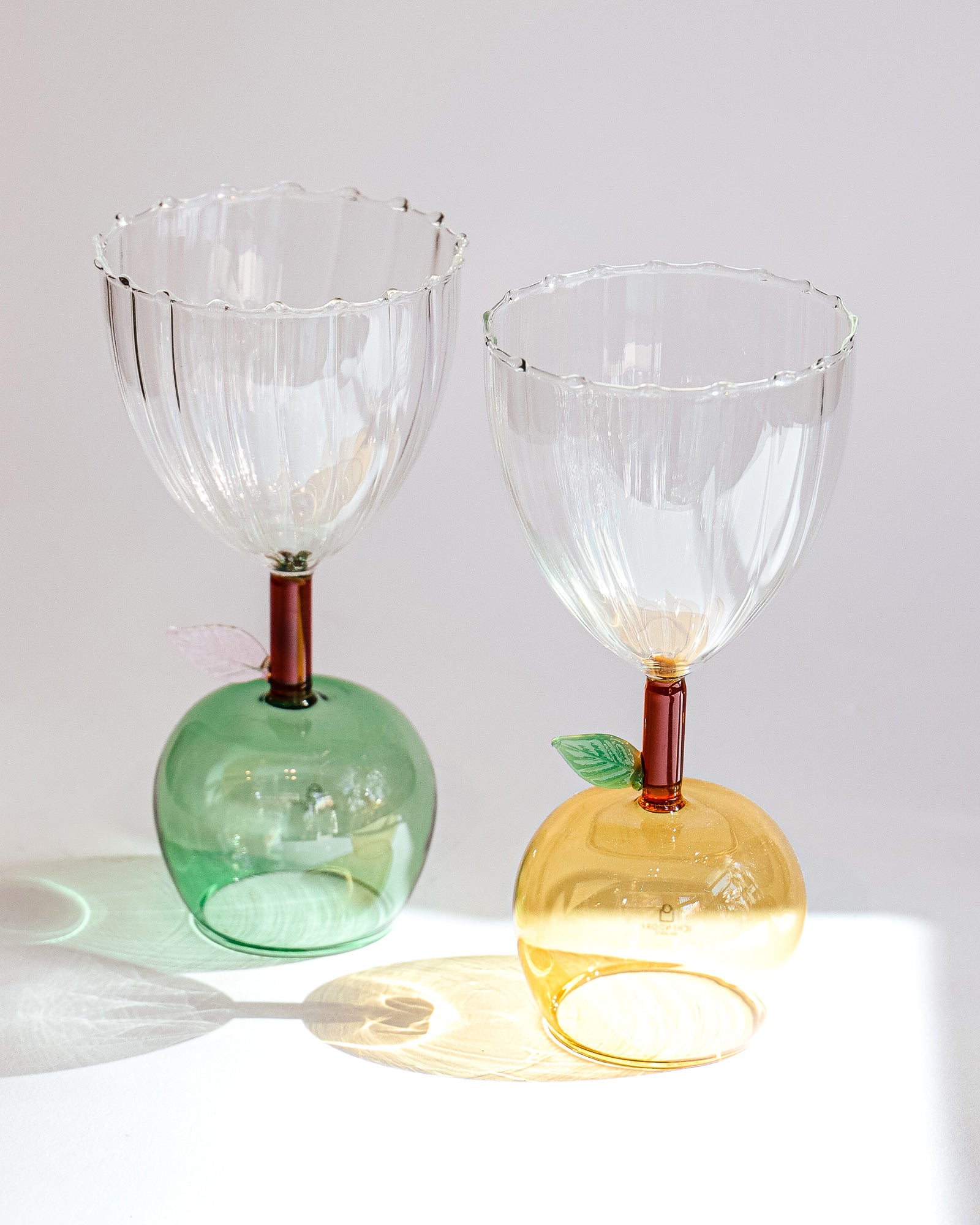 Two apple-shaped wine glasses from Ichendorf Milano's Fruit and Flower collection, with clear bowls, yellow and green glass bases, and a delicate pink and green glass leaf detail. Shown on a white background and available at Wildschut Antiques.