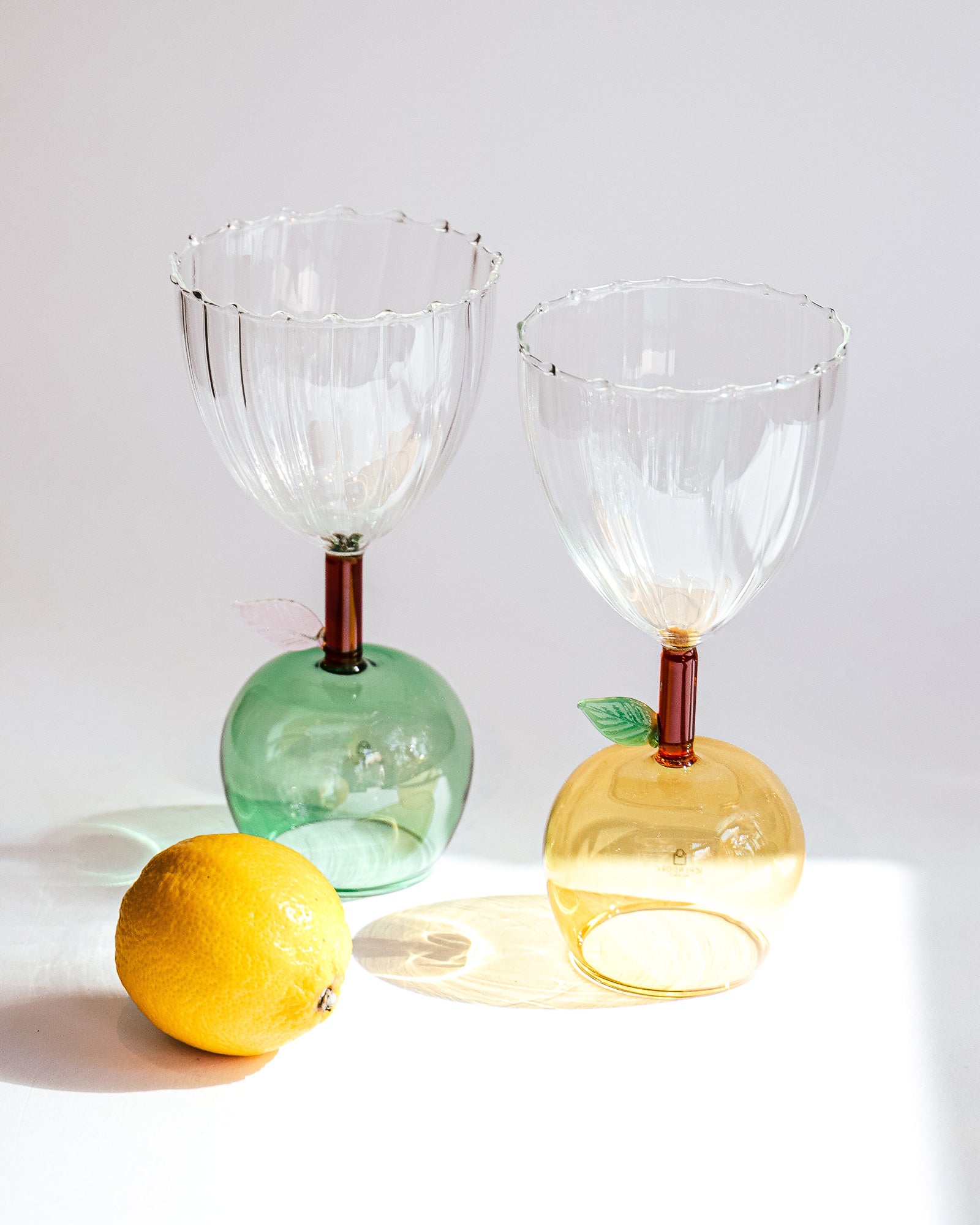 Two apple-shaped wine glasses from Ichendorf Milano's Fruit and Flower collection, with clear bowls, yellow and green glass bases, and a delicate pink and green glass leaf detail. Shown alongside a lemon on a white background and available at Wildschut Antiques.