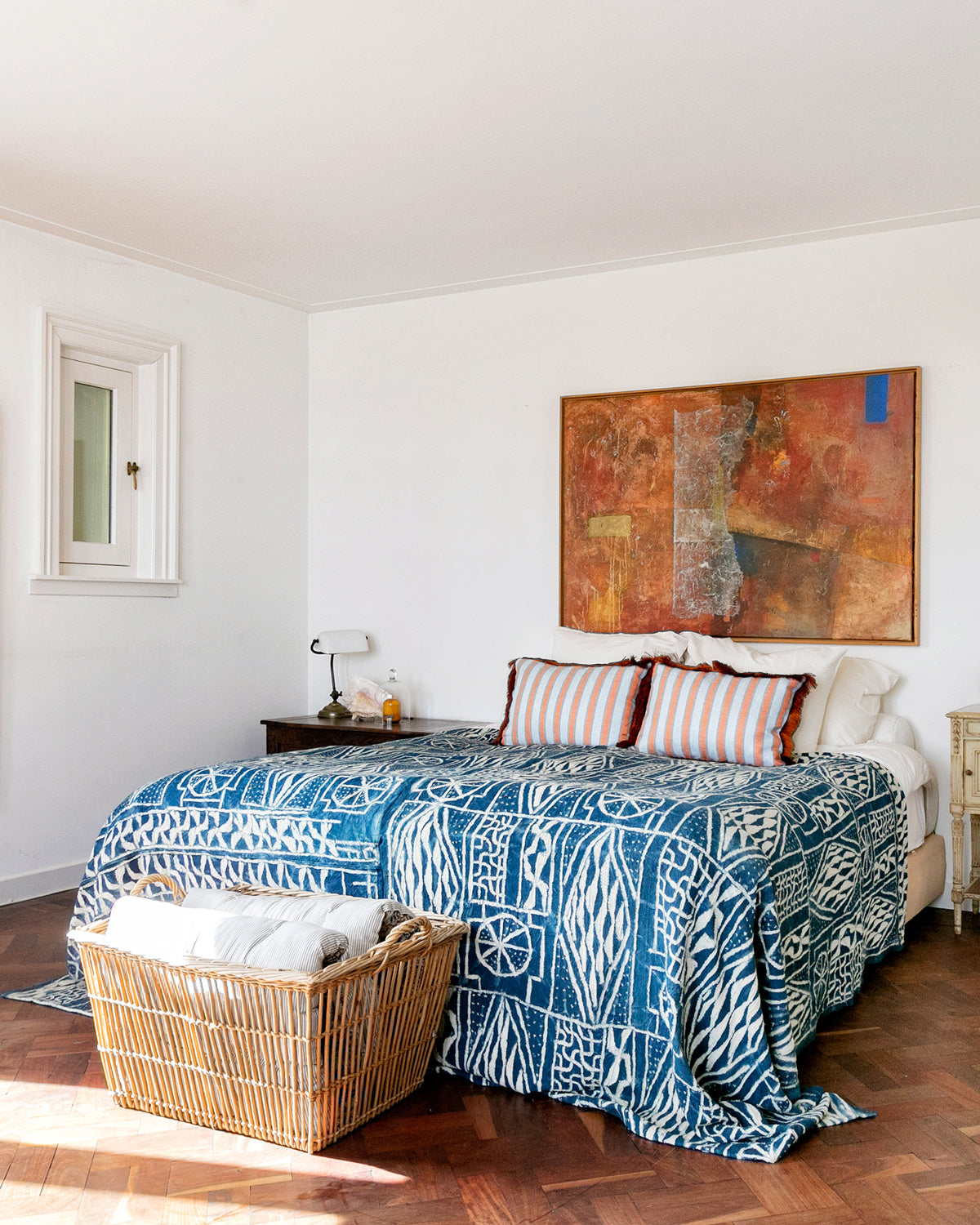 A vintage indigo-dyed 'Ndop' ceremonial textile with white geometric patterns, made from hand-spun cotton and raffia, from Cameroon, displayed on a large bed in a room with a wooden floor and a orange painting hanging above the bed