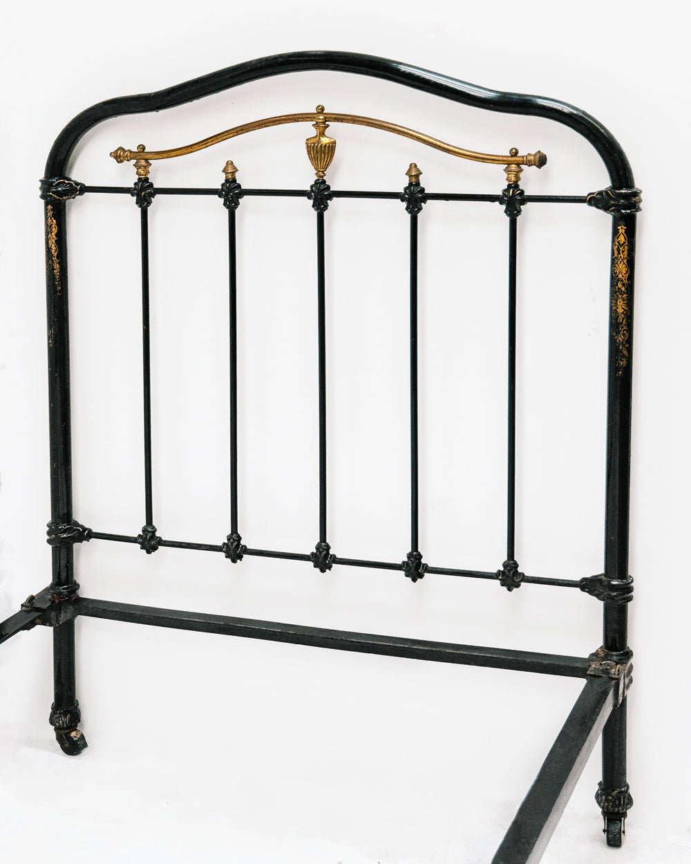Close-up of an antique cast iron single bed with black and gold finish, solid frame with intricate designs, small wheels on legs, no mattresses or slats included