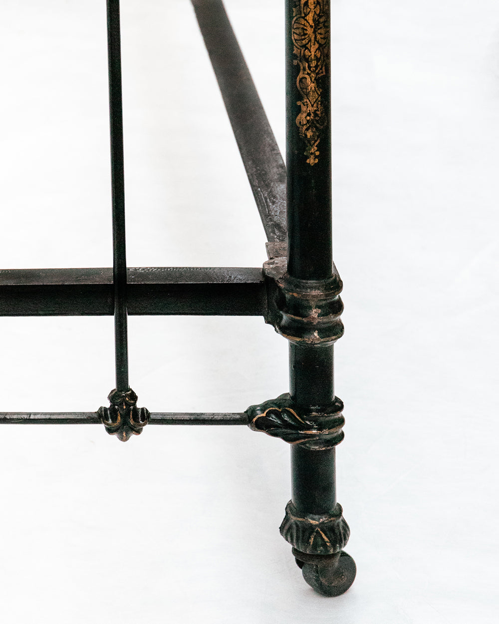 Close-up of an antique cast iron single bed with black and gold finish, solid frame with intricate designs, small wheels on legs, no mattresses or slats included