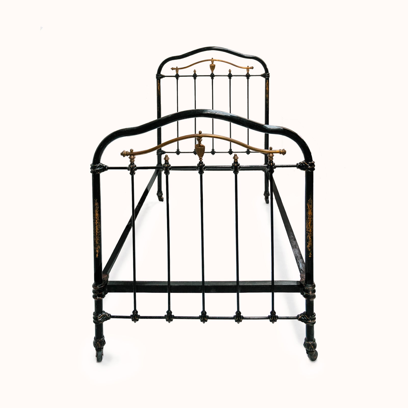 Antique cast iron single bed with black and gold finish, solid frame with intricate designs, small wheels on legs, no mattresses or slats included, available at Wildschut Antiques
