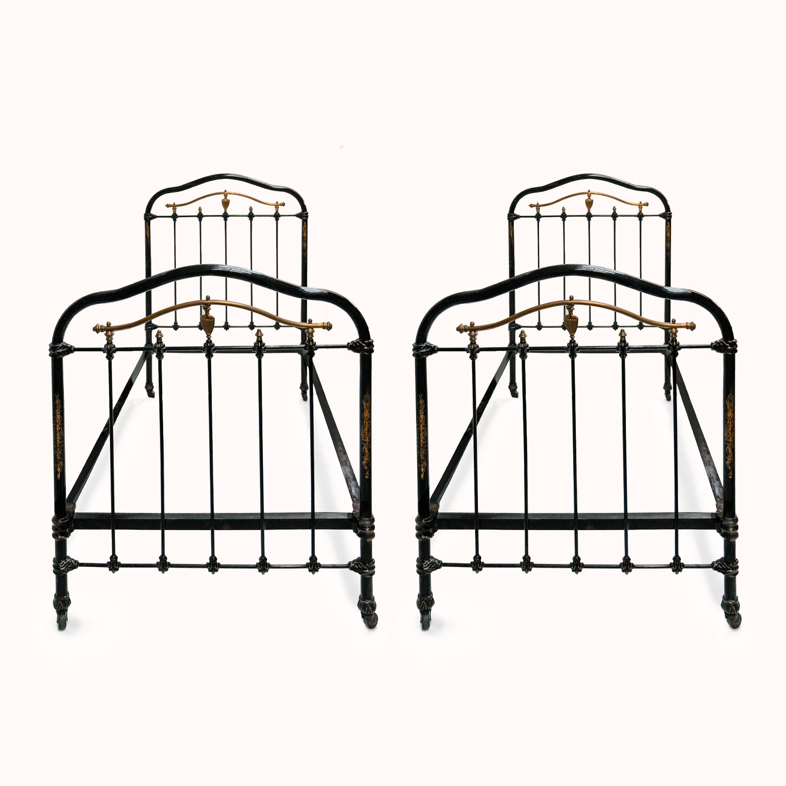Matching pair of antique cast iron single beds with black and gold finish, solid frames with intricate designs, small wheels on legs, no mattresses or slats included, available at Wildschut Antiques