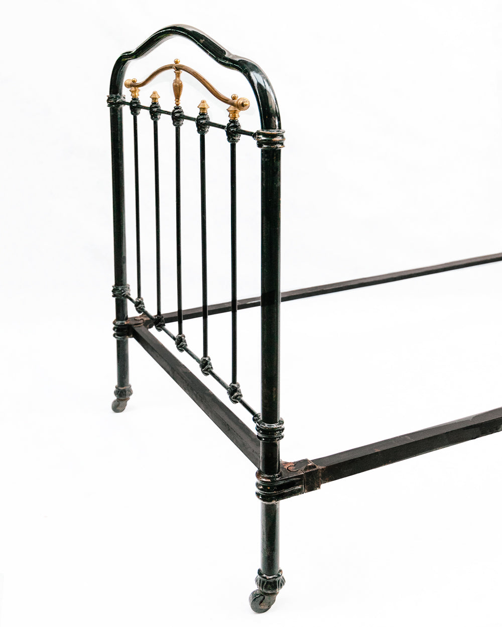 Close-up of an antique cast iron single bed with black and gold finish, solid frame with intricate designs, small wheels on legs, no mattresses or slats included