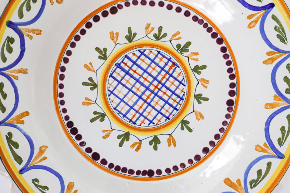 Detail of a small, vintage, hand-painted majolica ceramic plate with a geometric pattern, featuring swirls and foliate motifs in a combination of blue, green, and orange colors on a white background