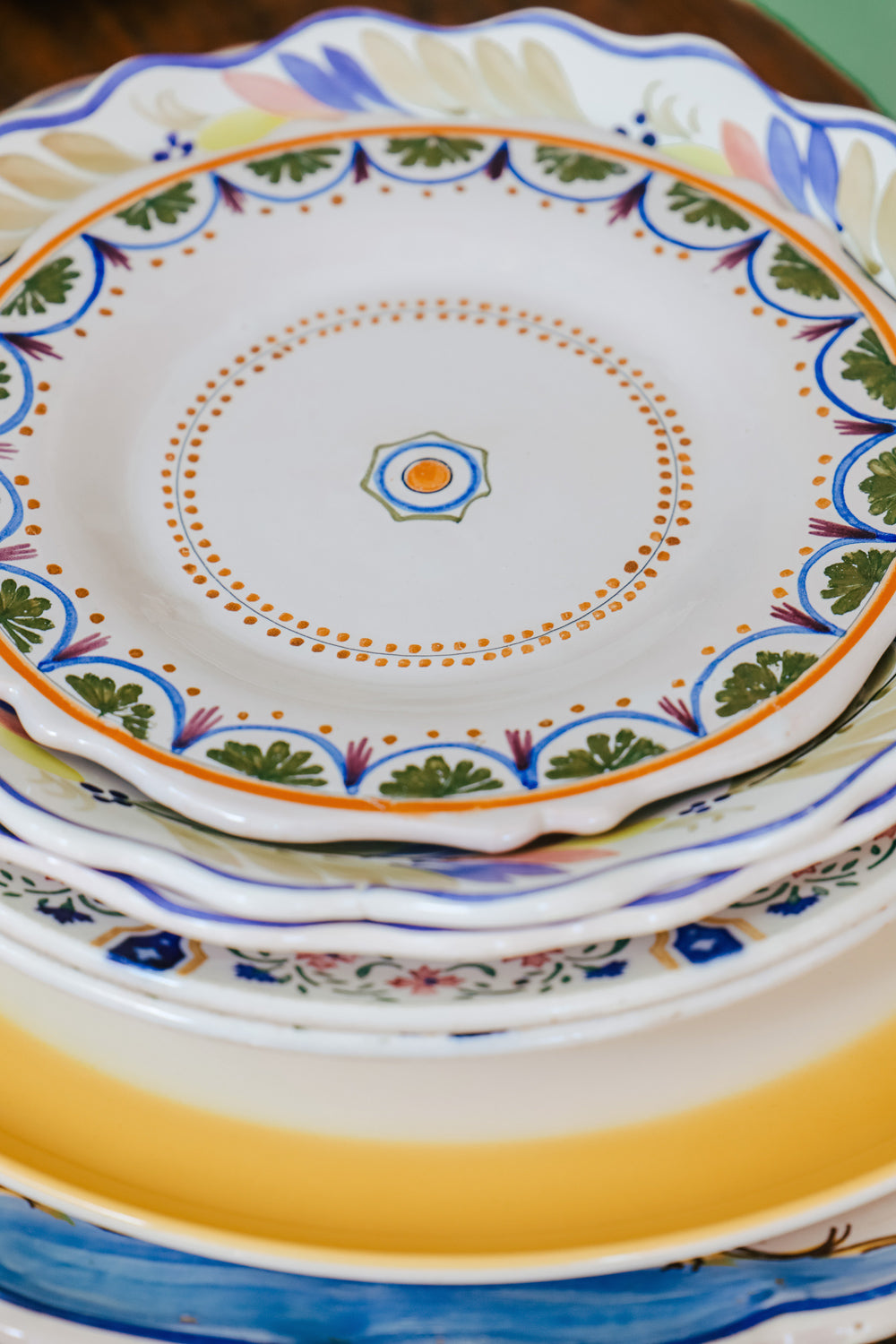 Stack of vintage ceramic plates crowned by a small hand-painted majolica plate showcasing a geometric design with swirls and foliate motifs in blue, green, and orange on a white background, available at Wildschut Antiques.