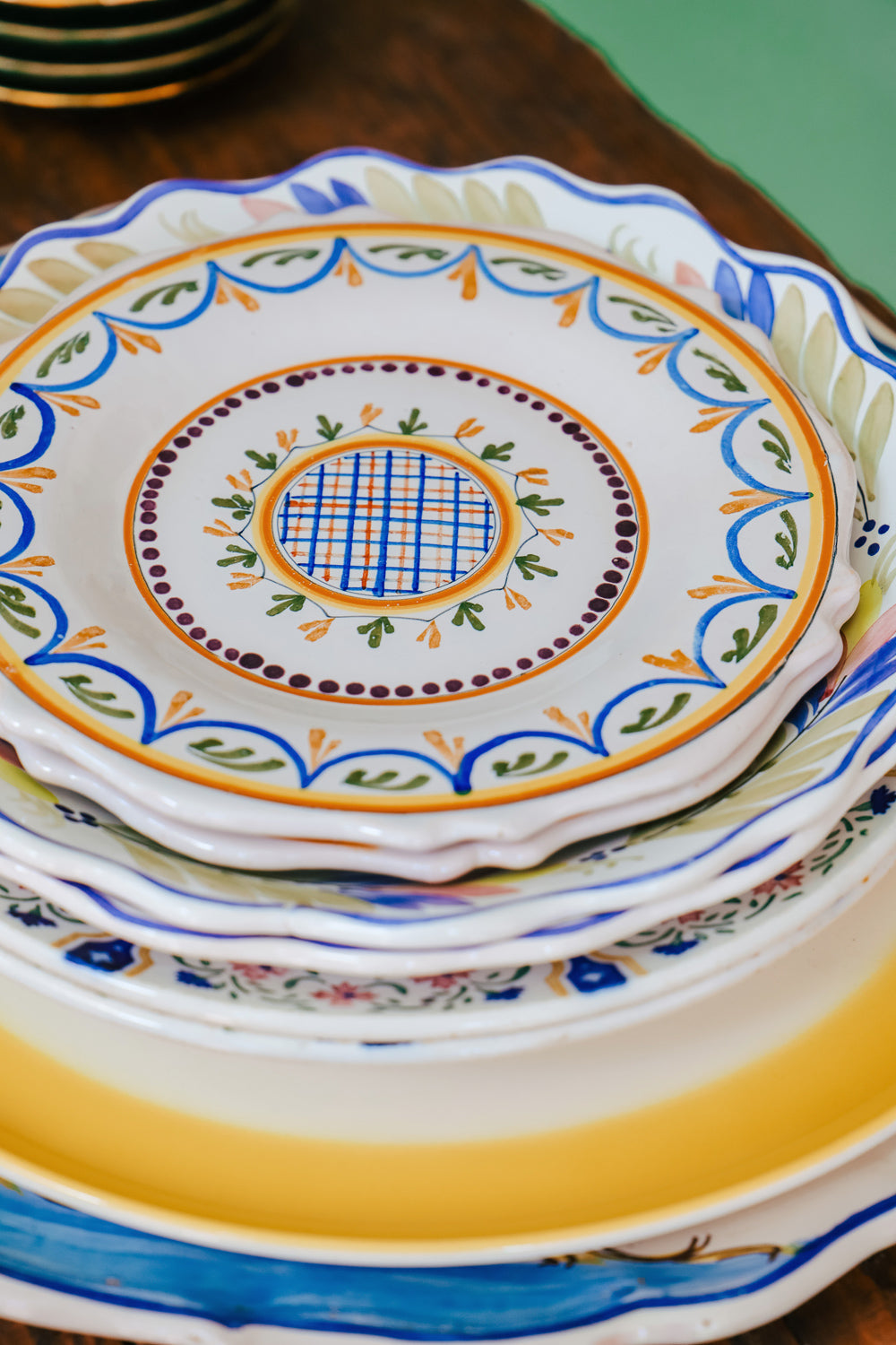 Stack of vintage ceramic plates crowned by a small hand-painted majolica plate showcasing a geometric design with swirls and foliate motifs in blue, green, and orange on a white background, available at Wildschut Antiques.