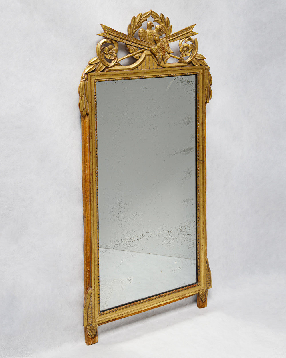 Sideview of an antique French Louis XVI period giltwood trumeau marriage mirror with a detailed crest featuring two birds and crossed arrows, surrounded by laurel garlands available at Wildschut Antiques