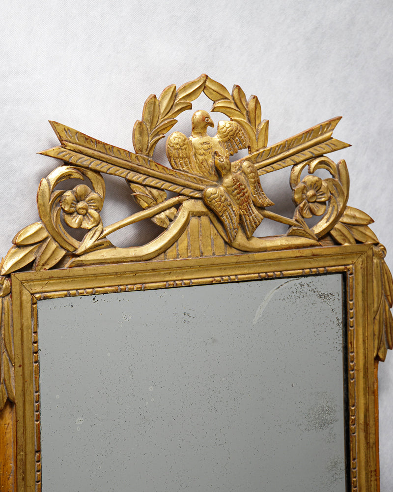 Close-up of an antique French Louis XVI period giltwood trumeau marriage mirror with a detailed crest featuring two birds and crossed arrows, surrounded by laurel garlands
