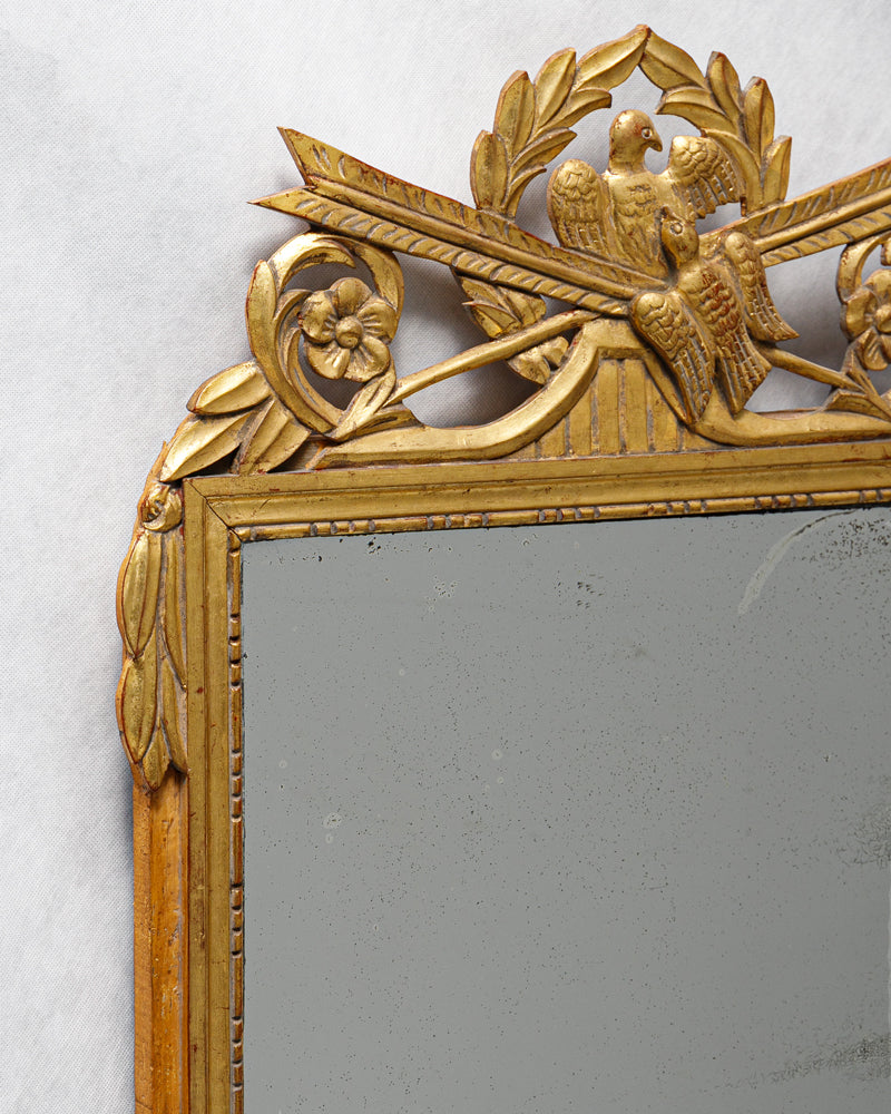 Close-up of an antique French Louis XVI period giltwood trumeau marriage mirror with a detailed crest featuring two birds and crossed arrows, surrounded by laurel garlands