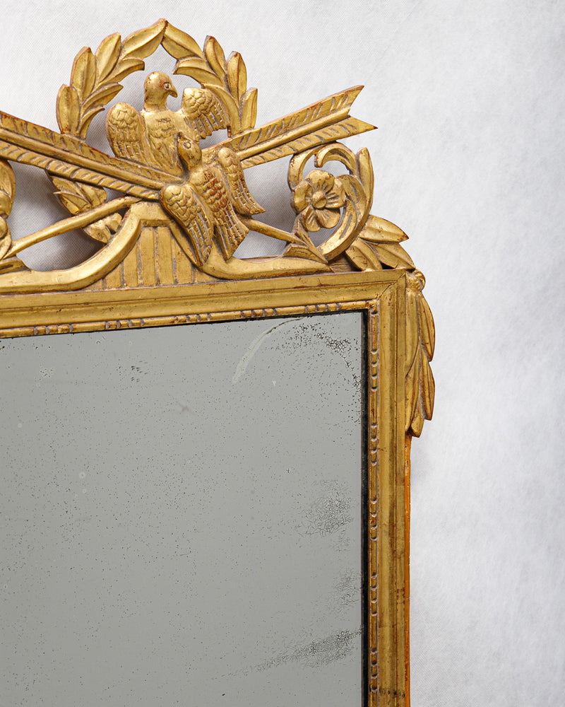 Close-up of an antique French Louis XVI period giltwood trumeau marriage mirror with a detailed crest featuring two birds and crossed arrows, surrounded by laurel garlands
