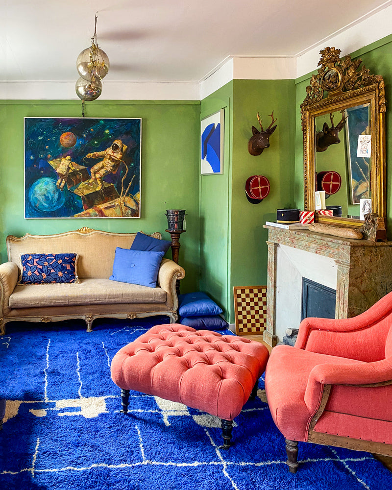 Colorful living room with green walls, a blue rug, and a pink sofa chair, an antique golden mirror, witch balls and an oil painting with astronauts