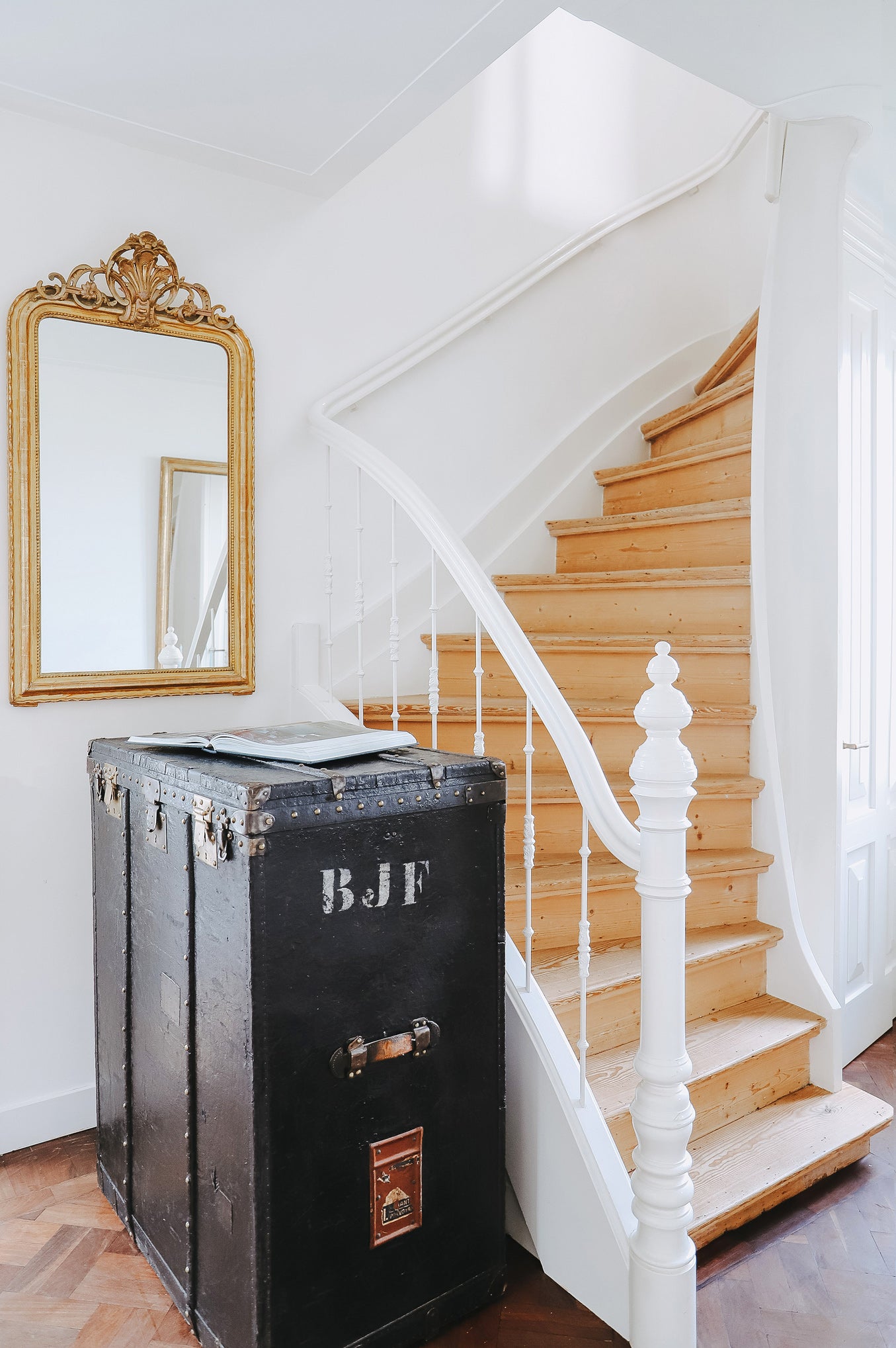 Antique 19th-century French Louis Philippe-style gilt mirror with decorative crest and pearl beading along the inner perimeter, displayed in a hallway with white walls, wooden stairs, and an antique black trunk.