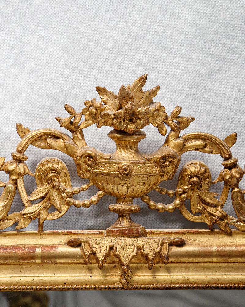 Close-up of an antique French gold gilt mirror with a vase and flower crest, featuring a classic 'string of pearls' motif and hints of red bole