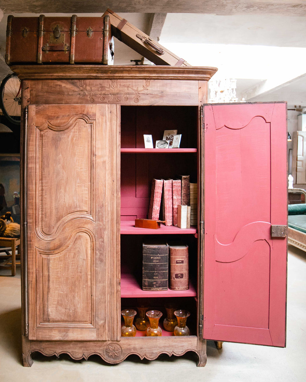 19th-century antique French Louis XV-style armoire with double doors—one open revealing a red painted interior filled with vases and old books, the other closed. Rustic wooden texture, suitcases stacked on top, photographed in the Wildschut Antiques warehouse.