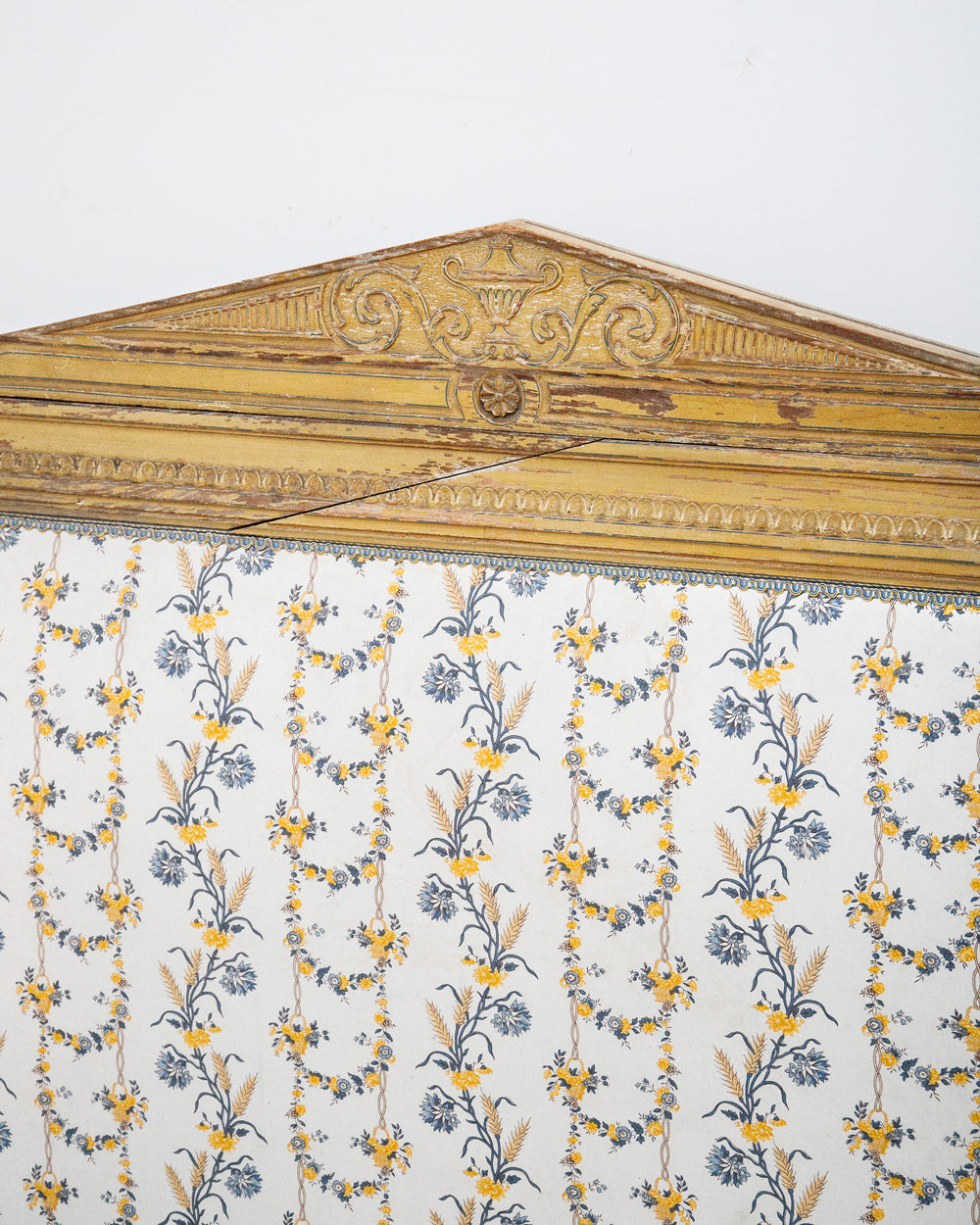 Close-up of an antique French hand-carved bed frame with a soft mustard yellow finish and floral upholstery in shades of white, yellow, and blue, in a neoclassical Louis XVI style