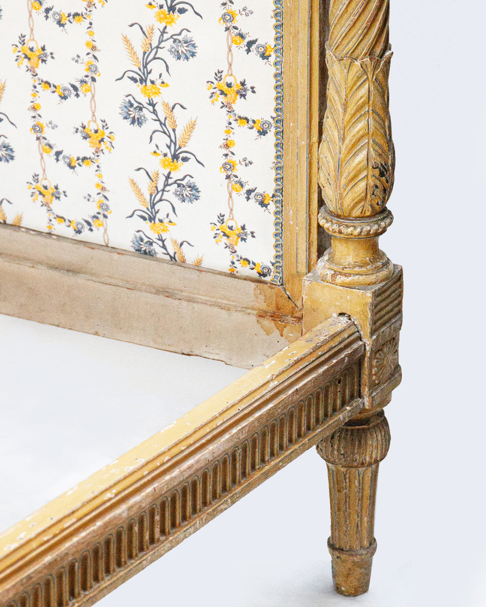Close-up of an antique French hand-carved bed frame with a soft mustard yellow finish and floral upholstery in shades of white, yellow, and blue, in a neoclassical Louis XVI style