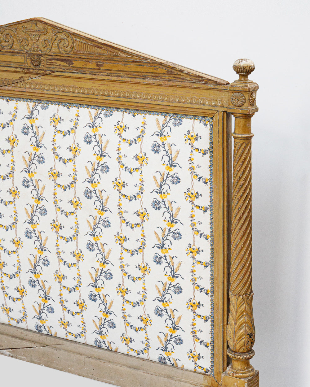 Close-up of an antique French hand-carved bed frame with a soft mustard yellow finish and floral upholstery in shades of white, yellow, and blue, in a neoclassical Louis XVI style