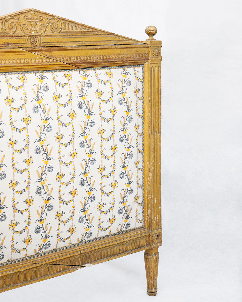 Close-up of an antique French hand-carved bed frame with a soft mustard yellow finish and floral upholstery in shades of white, yellow, and blue, in a neoclassical Louis XVI style