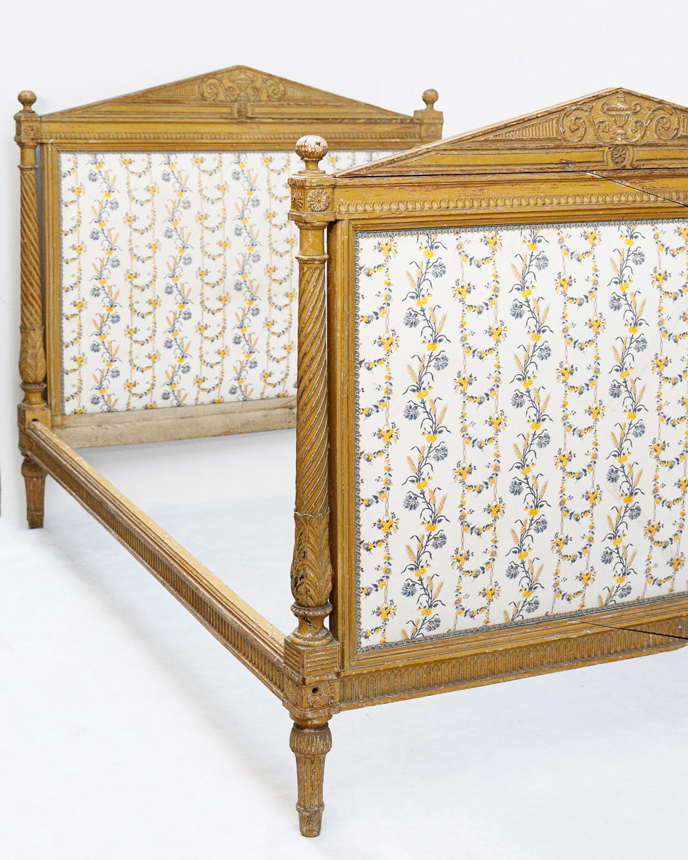 Close-up of an antique French hand-carved bed frame with a soft mustard yellow finish and floral upholstery in shades of white, yellow, and blue, in a neoclassical Louis XVI style