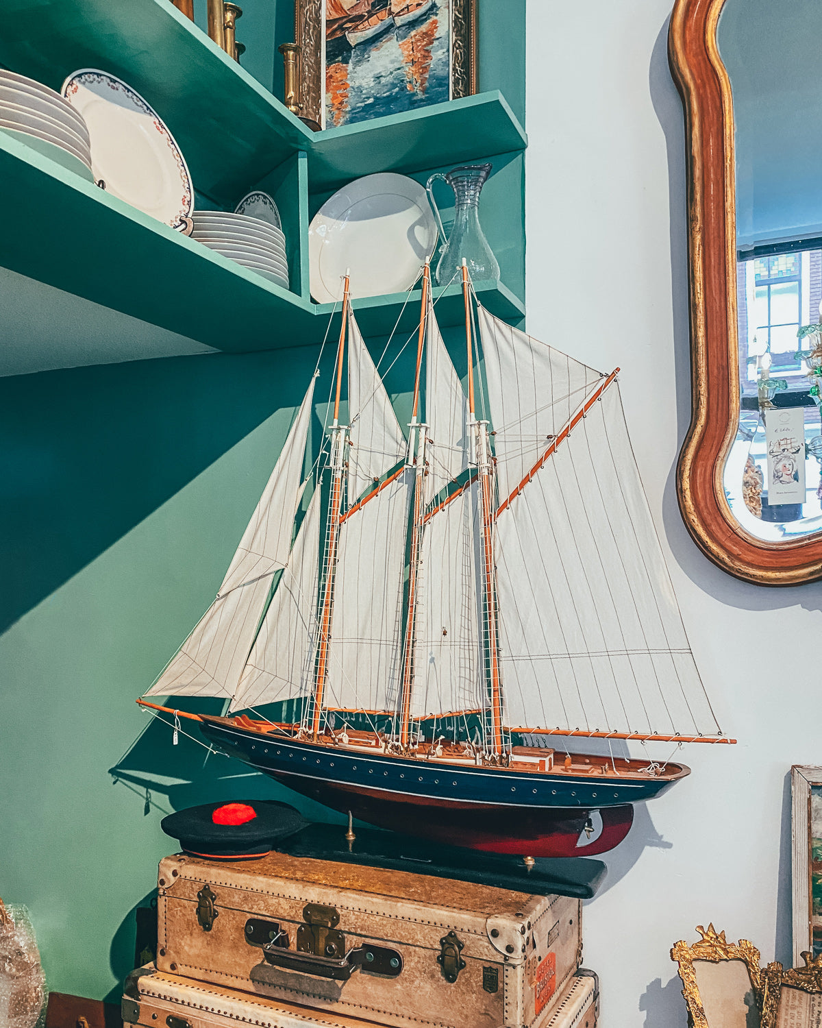 A vintage Marco Polo sail boat model with a predominantly white sail and a brown wooden hull, displayed on a stand, on top of a bunch of antique suitcases at Wildschut Antiques