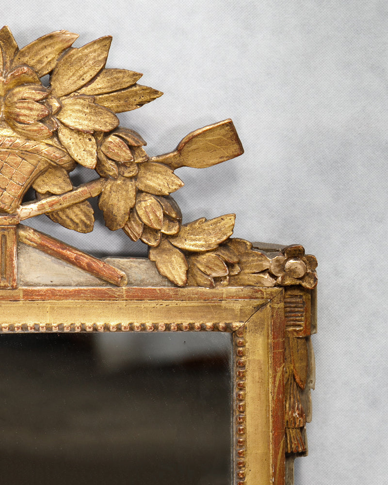 Close-up of an antique French Louis XVI period giltwood trumeau mirror with a finely carved crest featuring a flower-filled basket, laurel leaf garlands, tassels, and delicate pearl beading