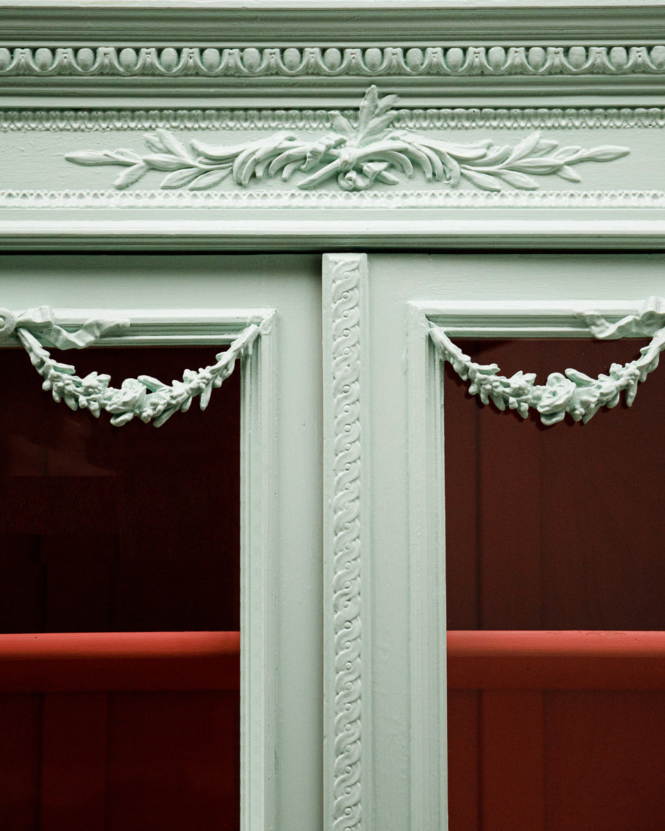 Close-up of a French Louis XVI-style vitrine cabinet with a soft mint green finish, glass doors, and contrasting red interior, intricate detailing.