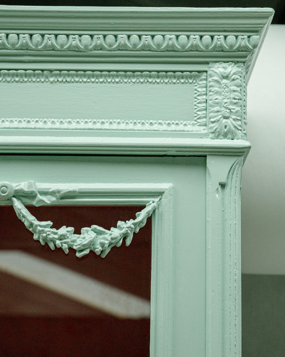 Close-up of the upper right corner of a French Louis XVI-style vitrine cabinet with a soft mint green finish, glass doors, and contrasting red interior, intricate detailing.
